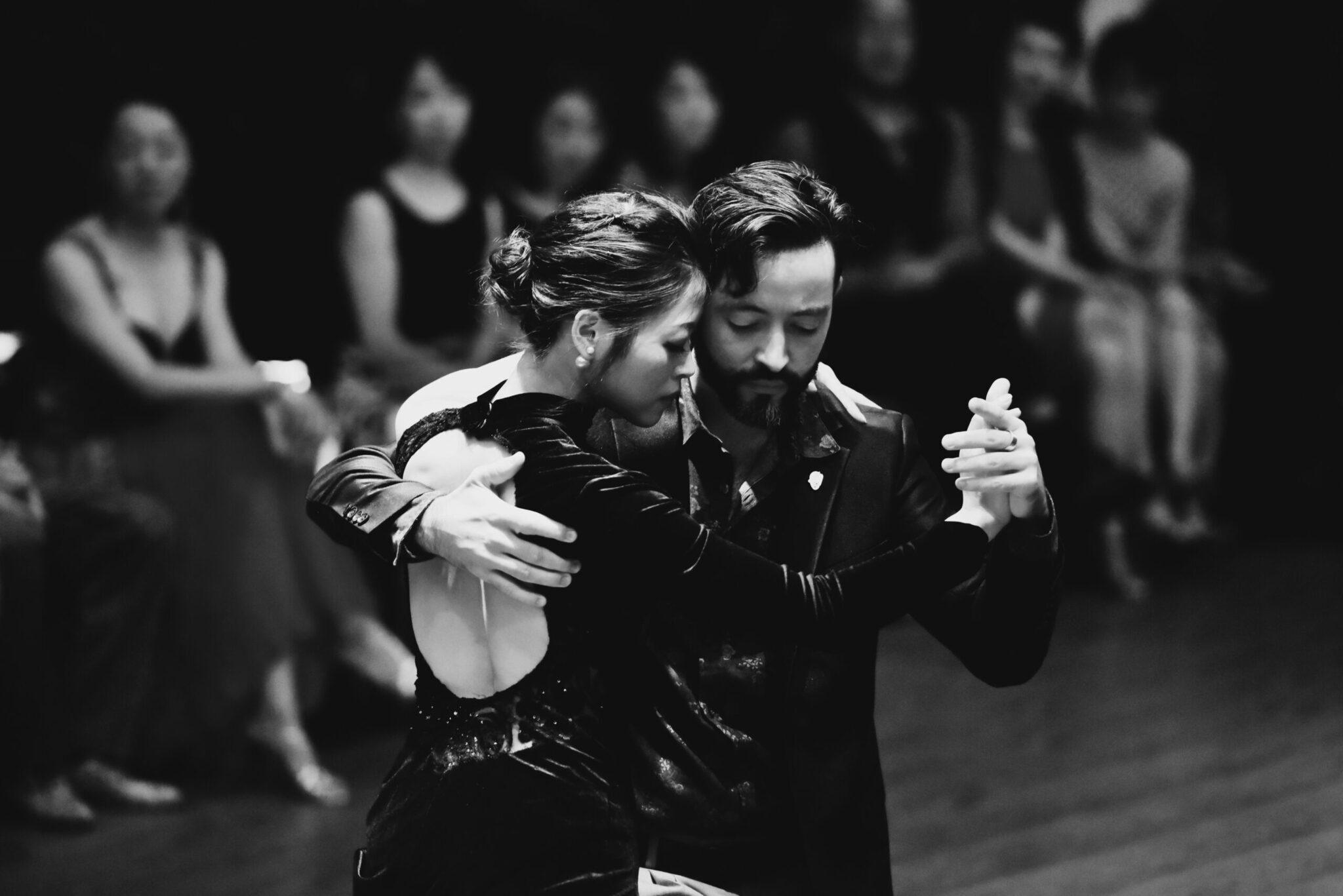 Coppia che danza il tango in costume nero davanti agli spettatori in un’atmosfera teatrale in bianco e nero