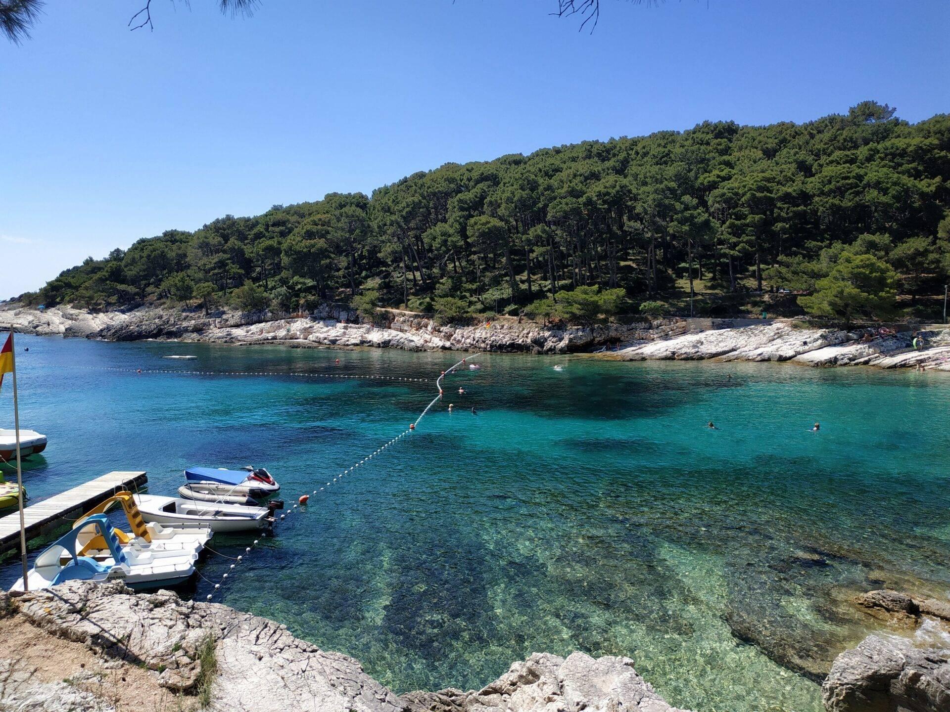 Una cala mediterranea con acque turchesi, barche ormeggiate, scogliere rocciose e foresta di pini