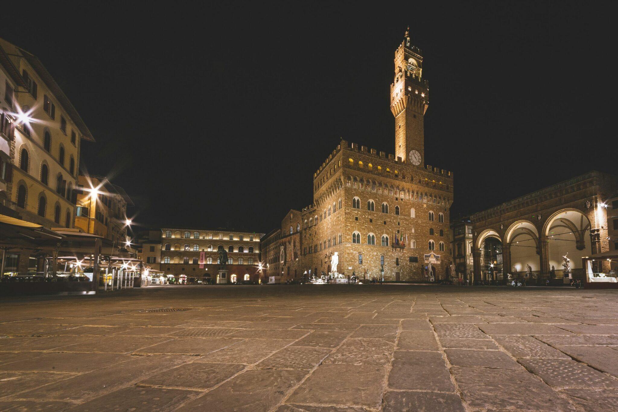 Plaza histórica italiana iluminada de noche con un palacio medieval coronado por una torre con reloj
