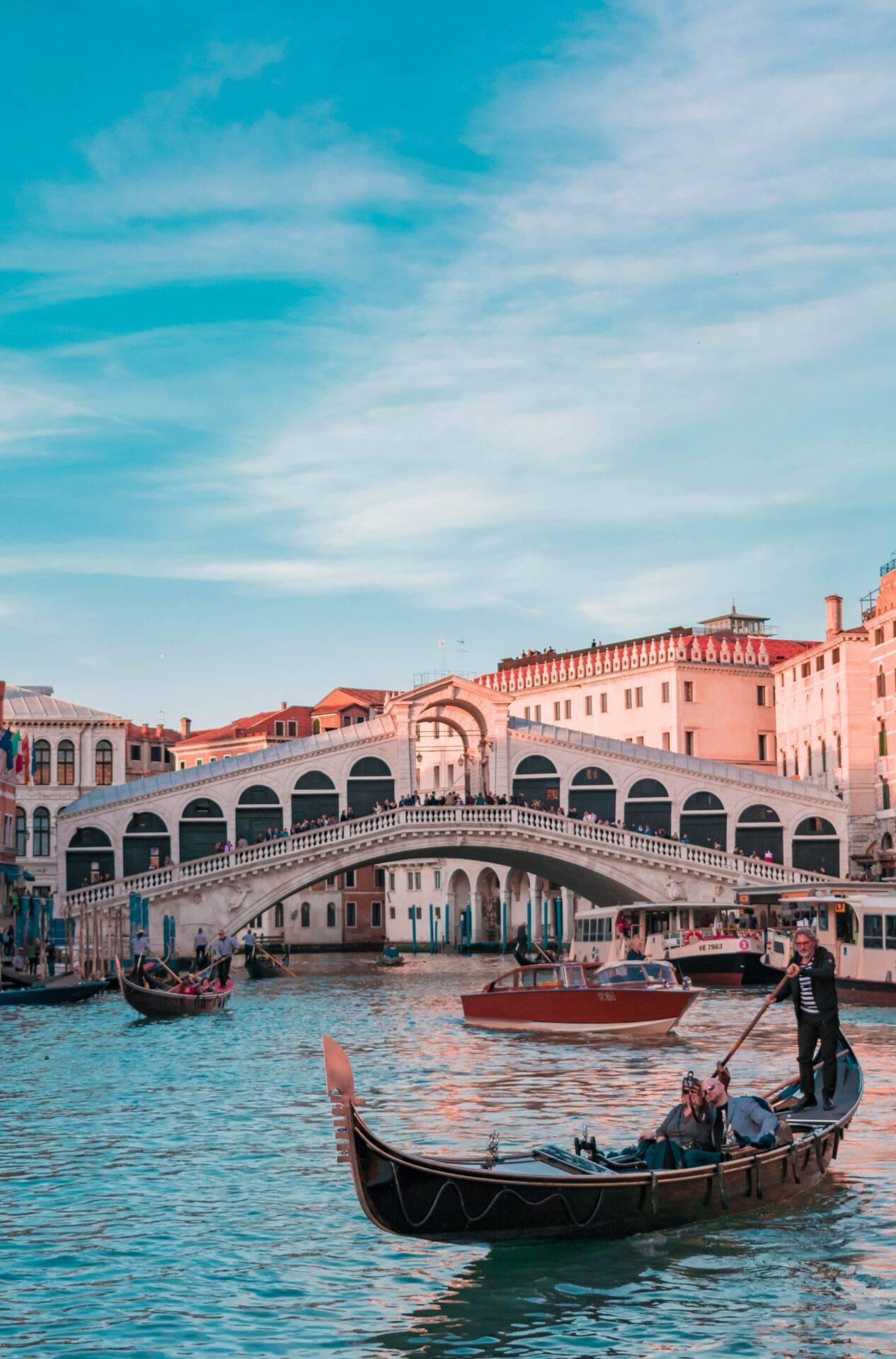 Gondole e turisti sul Canal Grande davanti al Ponte di Rialto con facciate rosa e architetture veneziane tipiche