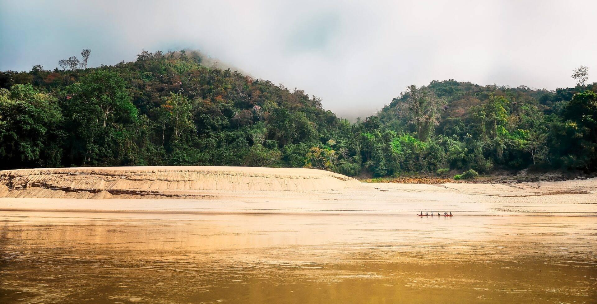 Una piccola barca tradizionale naviga su un fiume dalle acque fangose, incorniciata da rive sabbiose e foreste tropicali lussureggianti