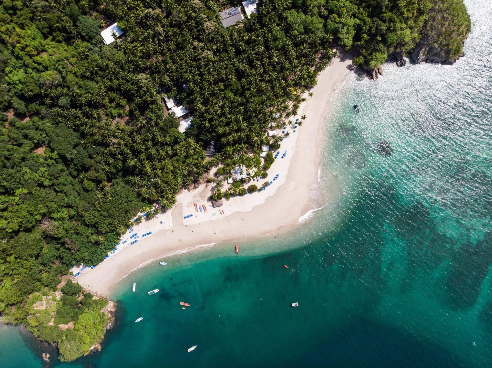 Luftaufnahme eines weißen Sandstrands, gesäumt von Regenwald, mit vor Anker liegenden Booten im kristallklaren türkisfarbenen Wasser