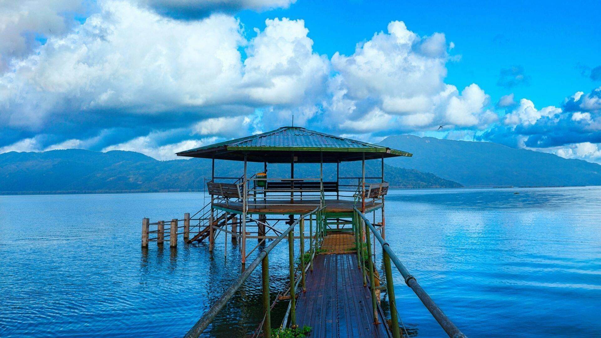 Un pontile in legno con riparo ombreggiato si estende su un lago turchese, montagne boscose sullo sfondo sotto un cielo nuvoloso