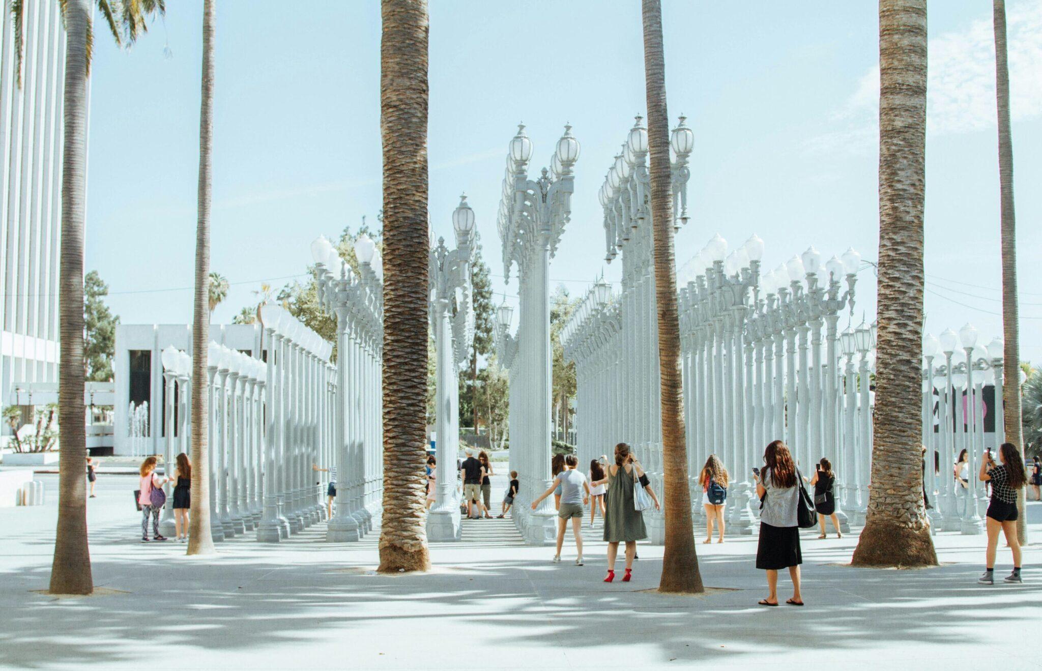 Una plaza pública moderna con columnas blancas, palmeras y farolas, animada por visitantes bajo un cielo despejado