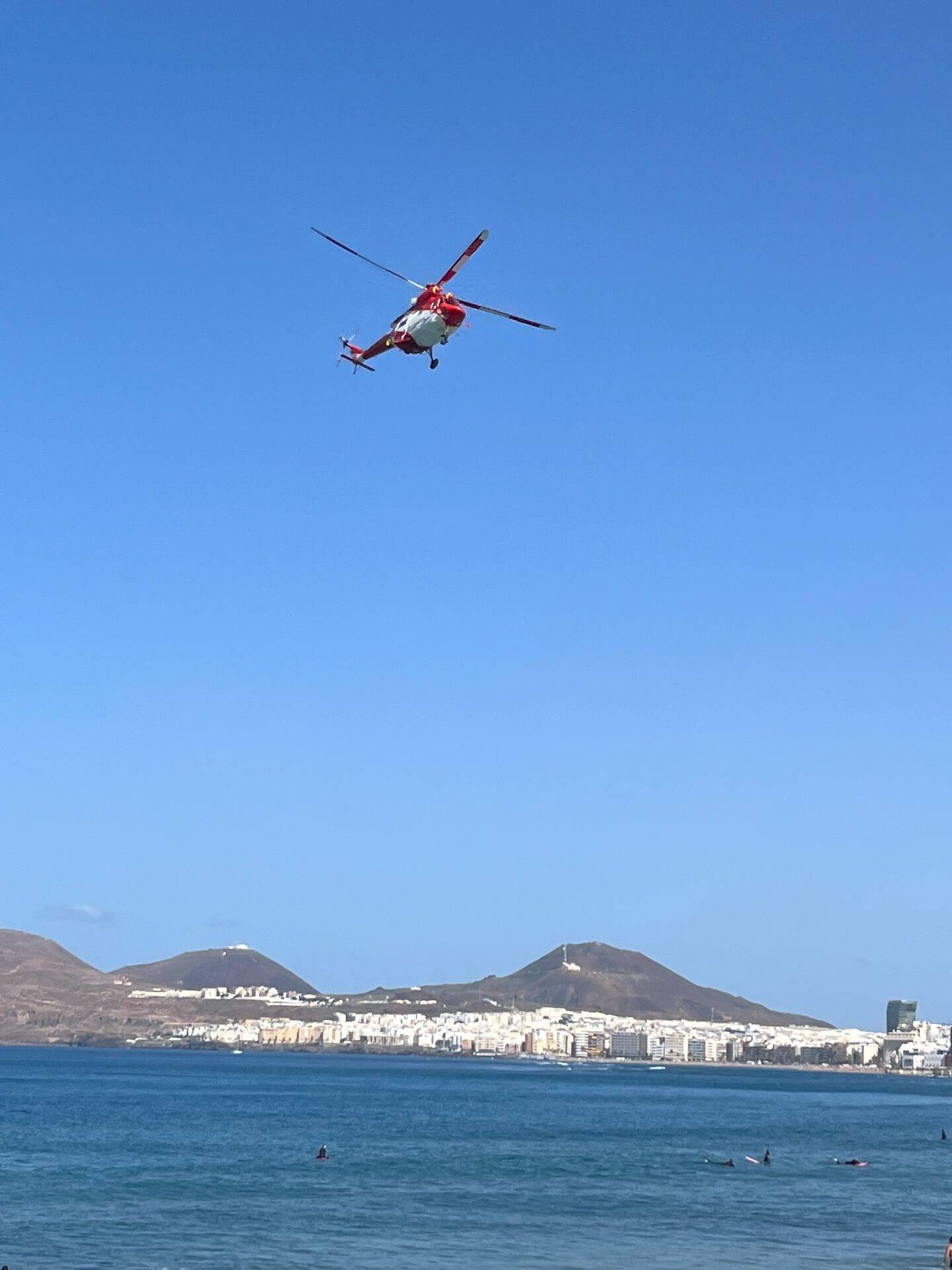 Un elicottero di soccorso sorvola una spiaggia con bagnanti di fronte a una città costiera e vulcani sullo sfondo