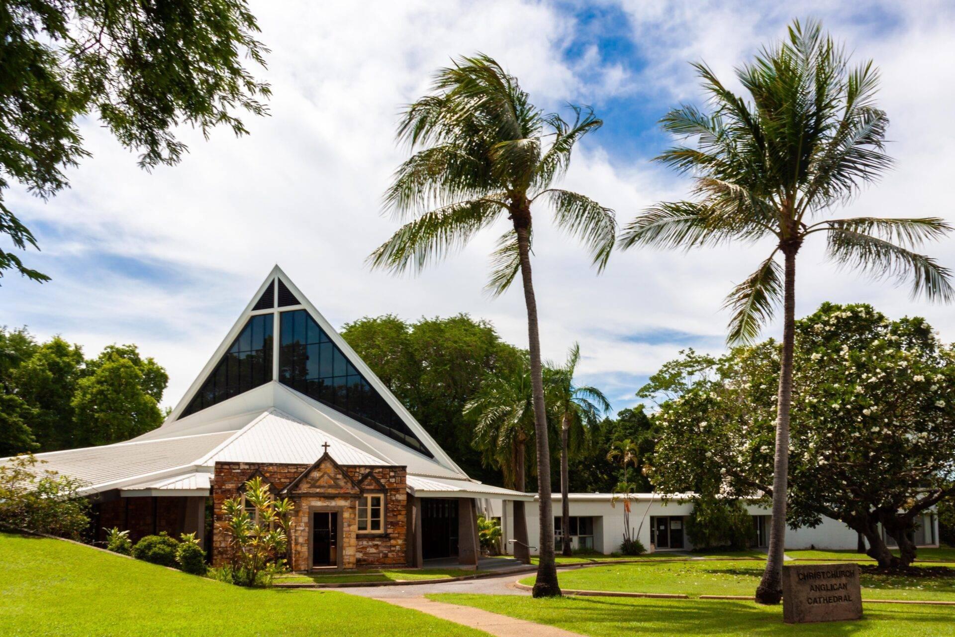 Una chiesa moderna con tetto bianco appuntito e croce, circondata da palme e verde sotto un cielo azzurro