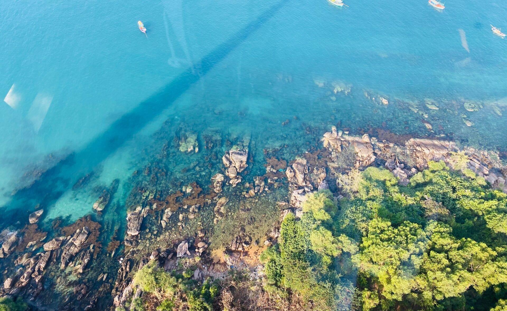 Vista aérea de una bahía turquesa con rocas costeras, vegetación verde y barcas de pesca amarradas