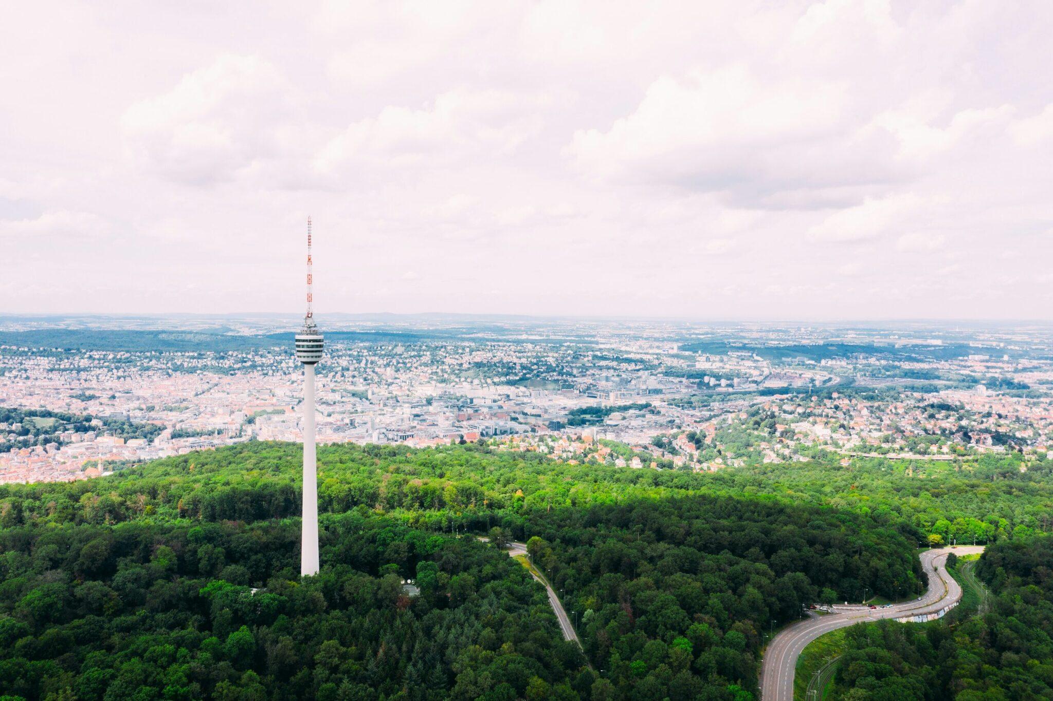 Vista aérea de una torre de televisión blanca que domina un denso bosque con una extensa ciudad al fondo