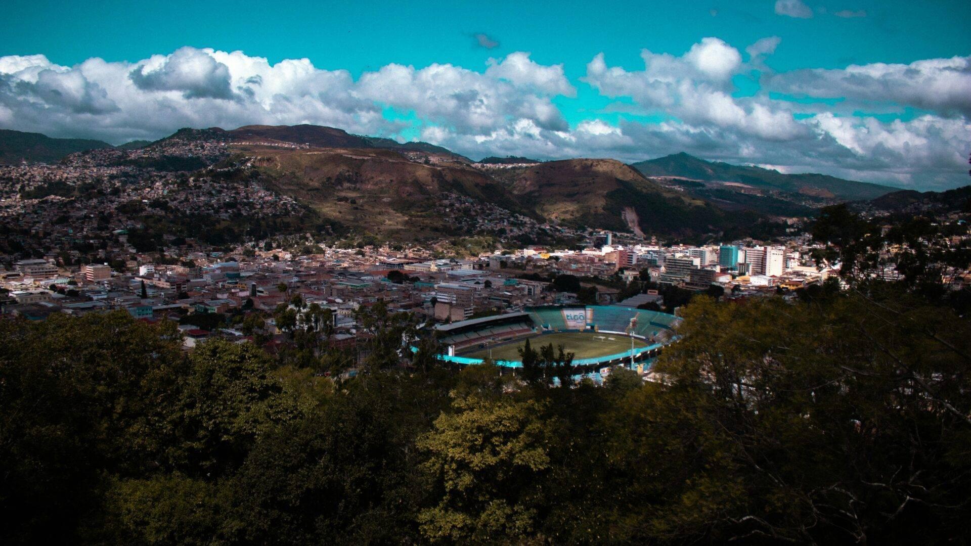 Vista panoramica di una città di montagna con uno stadio azzurro, circondata da colline boscose e edifici residenziali
