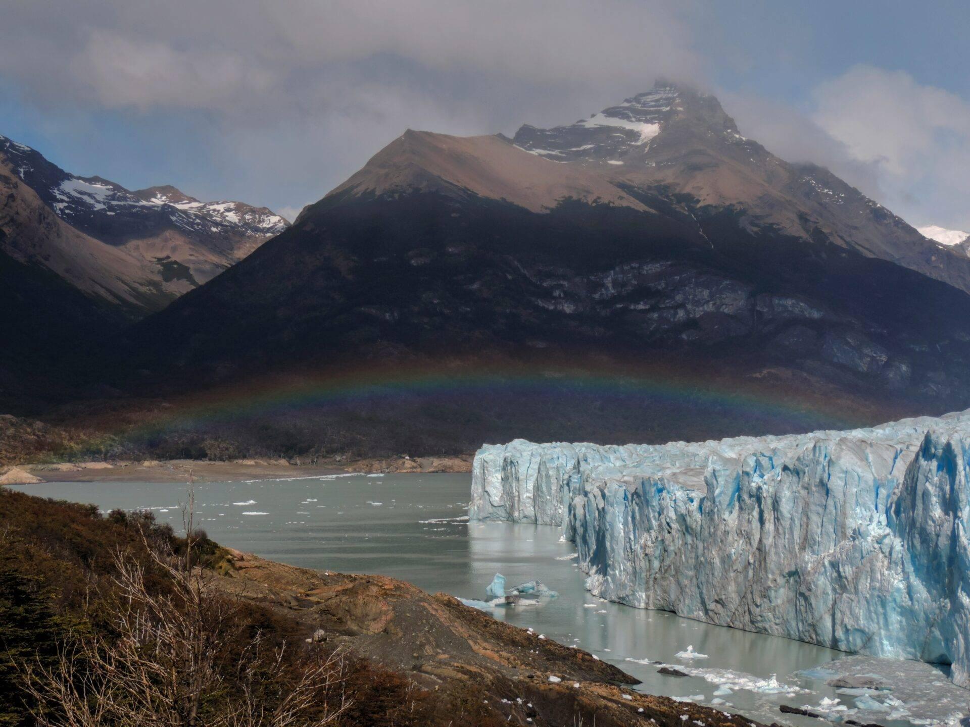 Weißer Gletscher gegenüber einem verschneiten Berg, der sich in einem türkisfarbenen Gletschersee spiegelt, mit Regenbogen