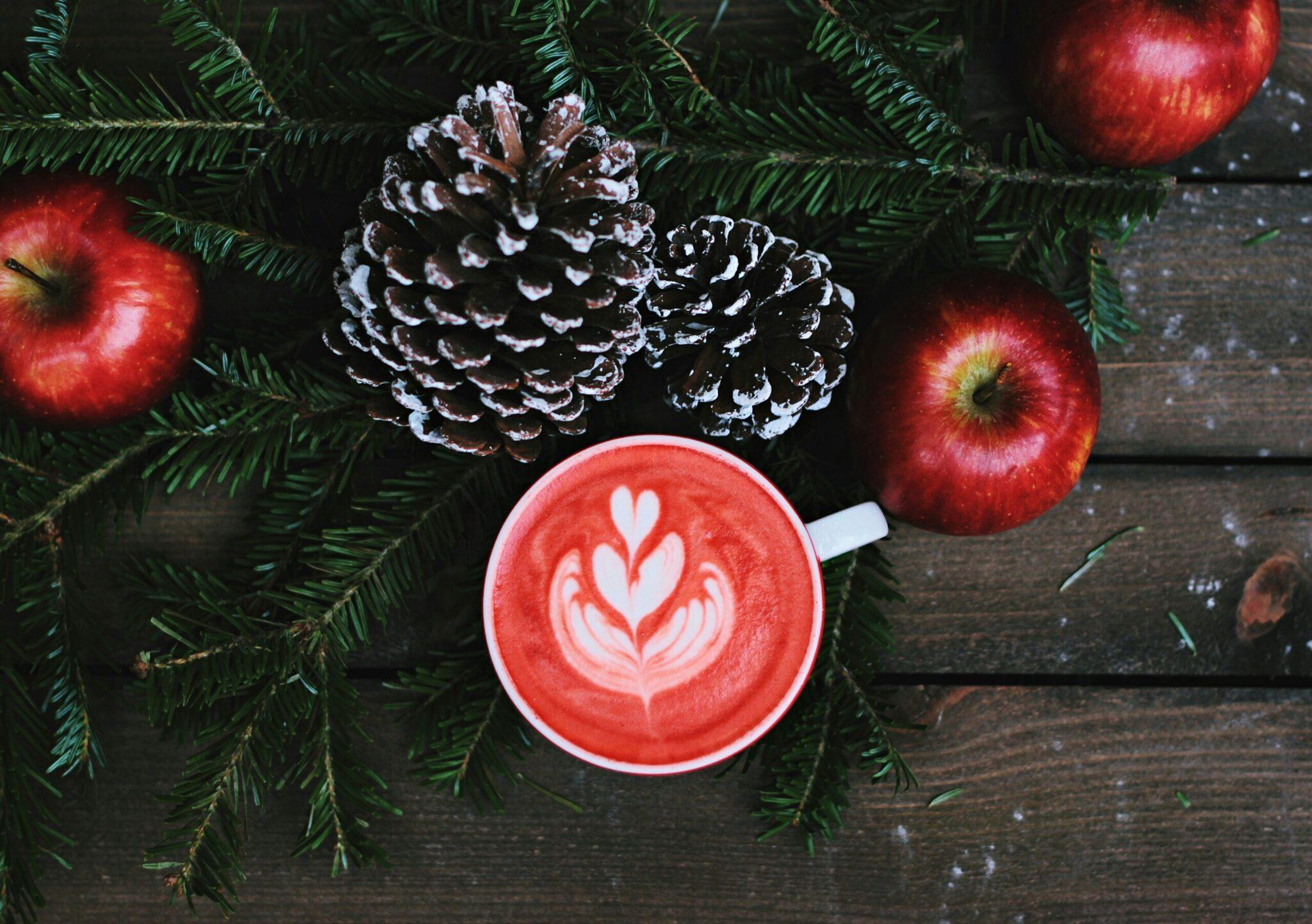 Taza de café con latte art en forma de corazón rodeada de ramas de abeto, manzanas rojas y piñas nevadas