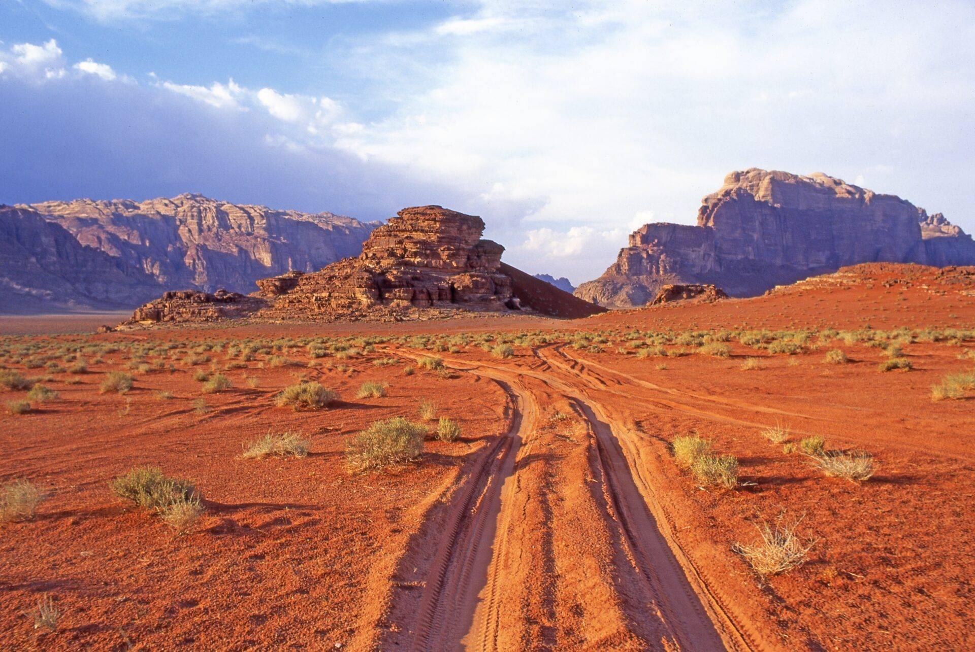 Camino de tierra roja que atraviesa un desierto árido con formaciones rocosas monumentales bajo un cielo azul con nubes