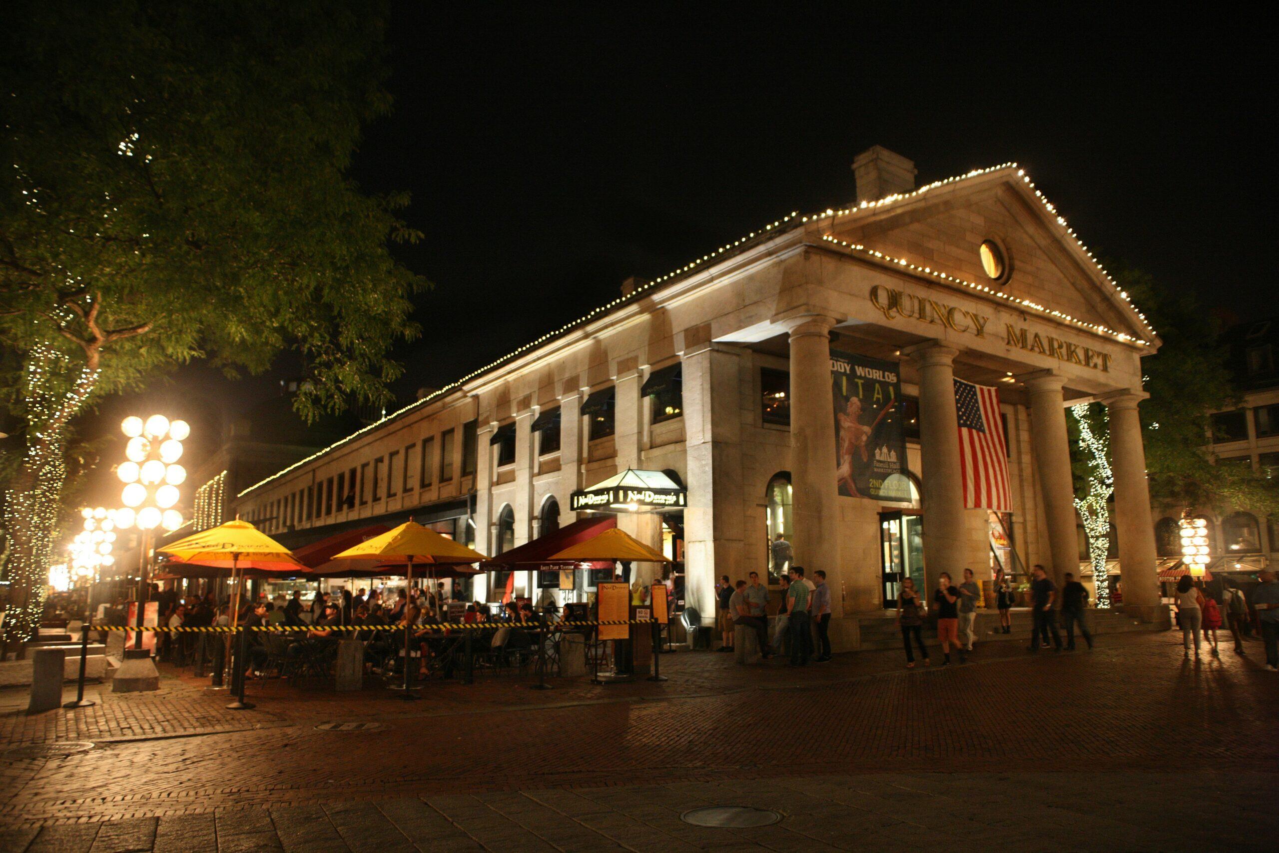The illuminated facade of a historic neoclassical market with columns, a terrace of parasols and evening strollers