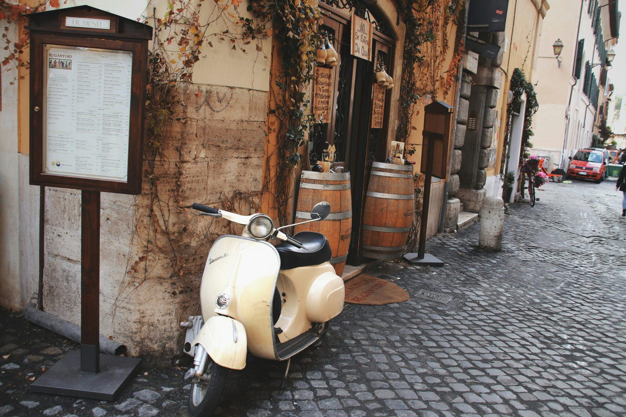 Un scooter Vespa blanco aparcado en un callejón empedrado de una ciudad europea antigua con fachadas ocres y hiedra