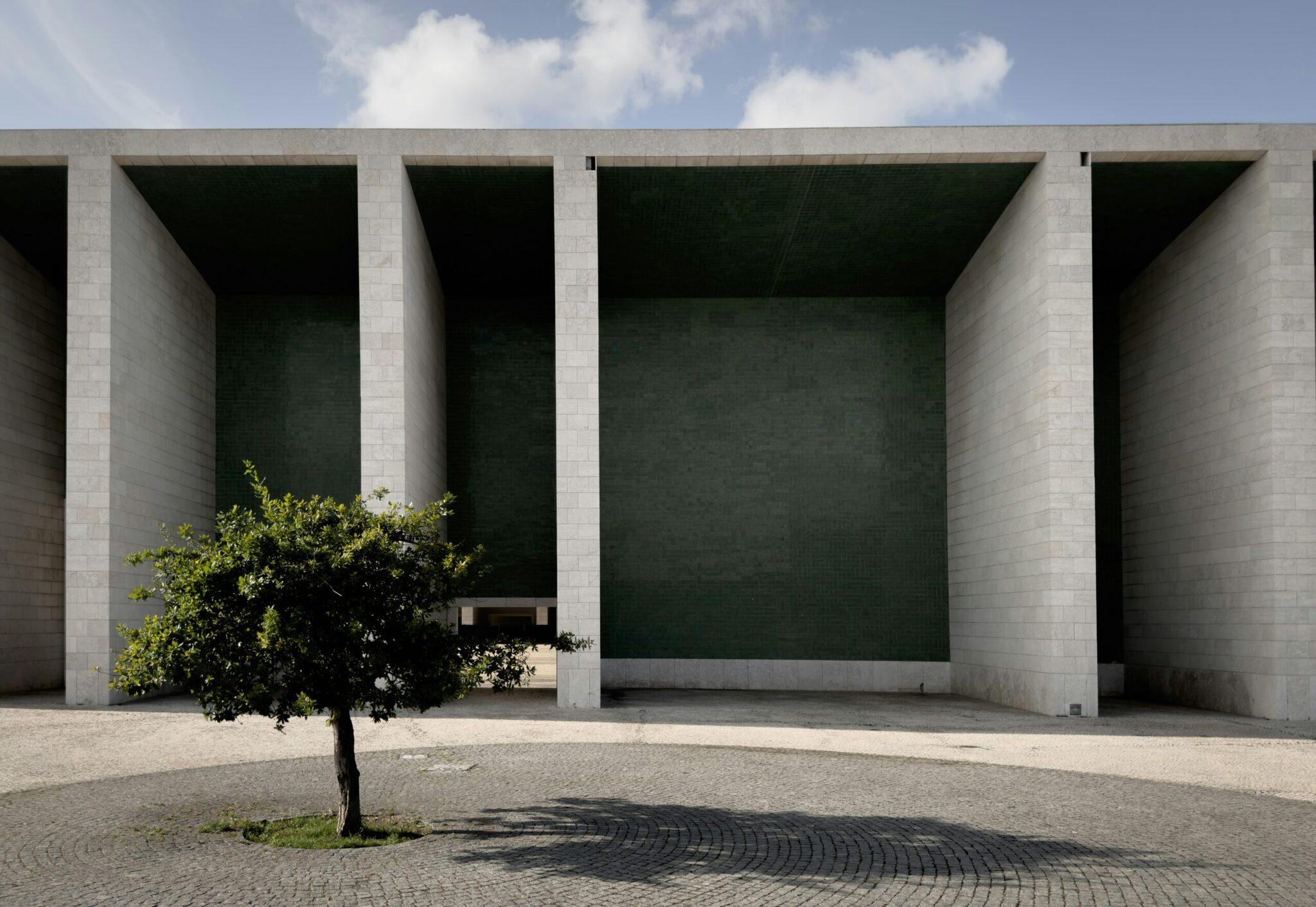 Moderne Betonarchitektur mit massiven Säulen, minimalistischem Innenhof und einzelnem Baum unter wolkenlosem Himmel