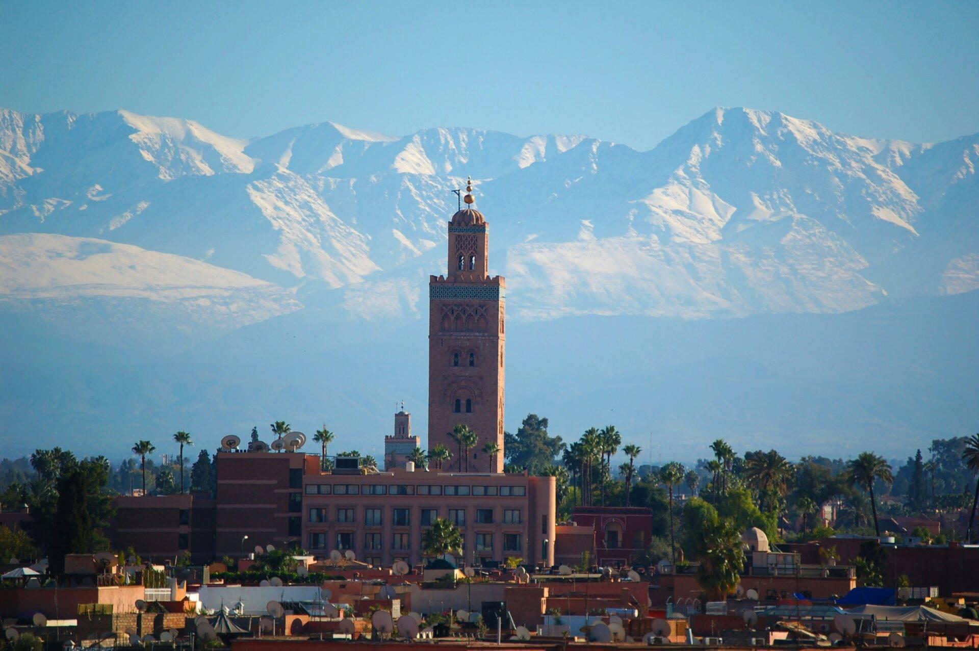 Minarete rojo ocre con decoraciones geométricas que domina la medina, montañas nevadas al fondo, palmeras y arquitectura tradicional