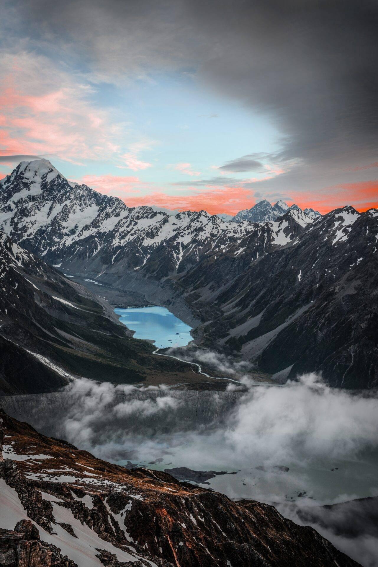 Catena montuosa innevata al tramonto con lago turchese incassato, valle nebbiosa e cielo rosato