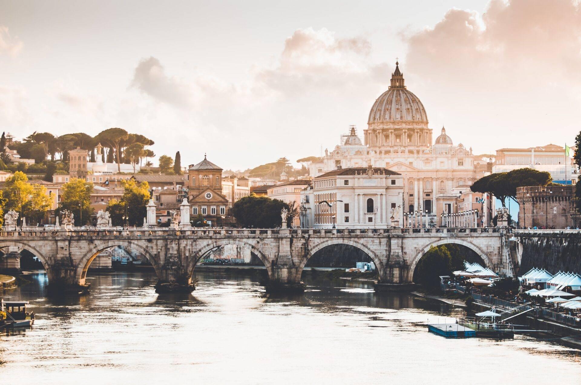 Panoramic view of a stone arch bridge spanning a river with a golden-domed basilica in the background