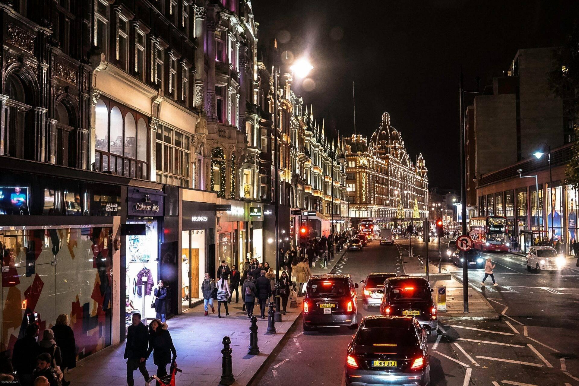 Una animada calle comercial de noche, engalanada para las fiestas, con fachadas victorianas, tiendas y taxis londinenses