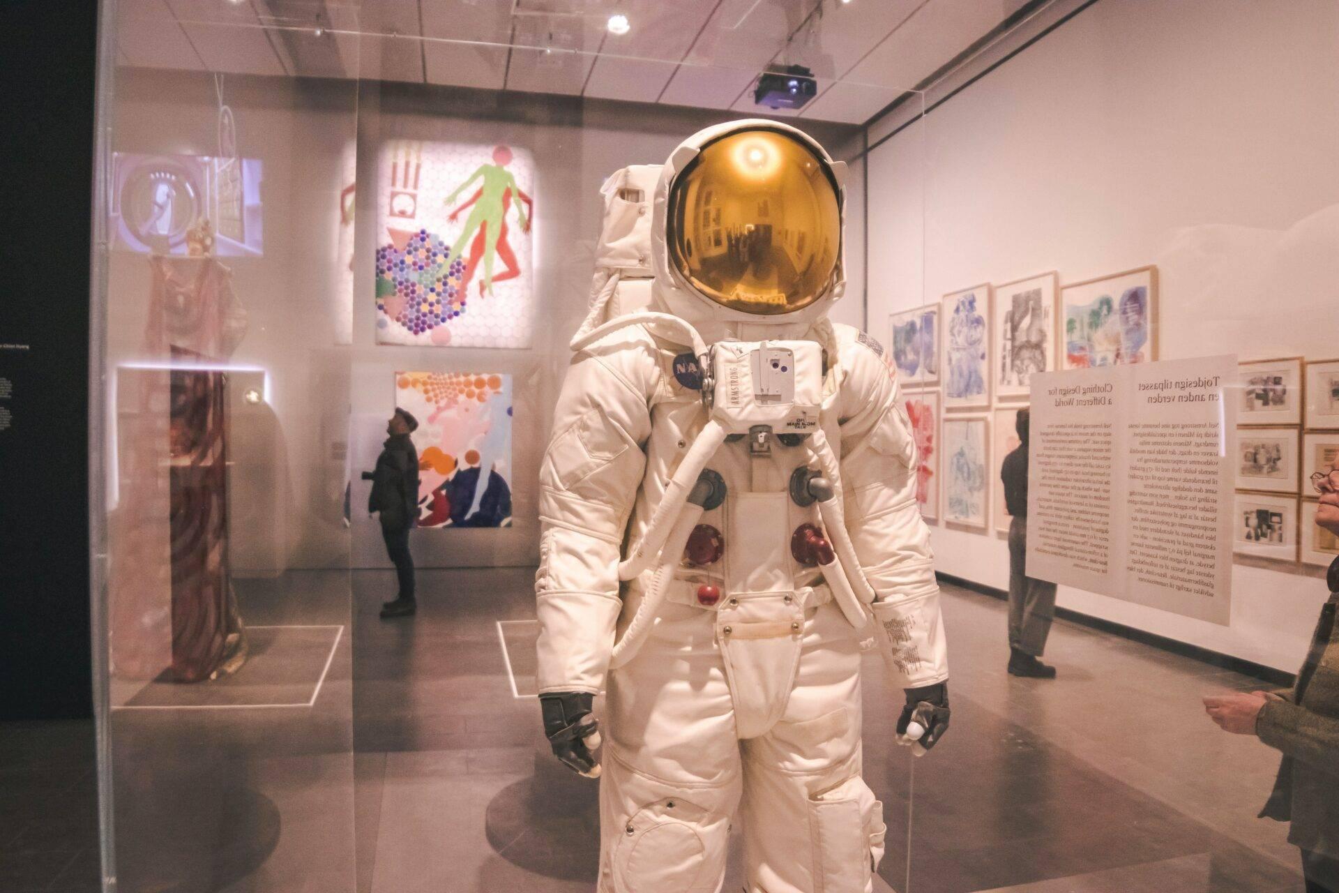 Una tuta spaziale bianca con casco dorato esposta in un museo, visitatori che osservano la mostra sullo sfondo