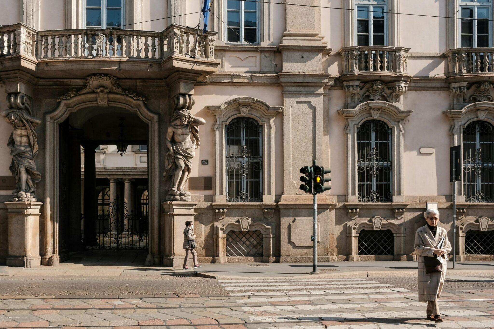 Ein gepflasterter Platz vor einem eleganten Renaissancepalast mit Säulen und Skulpturen, einige Fußgänger in der Sonne