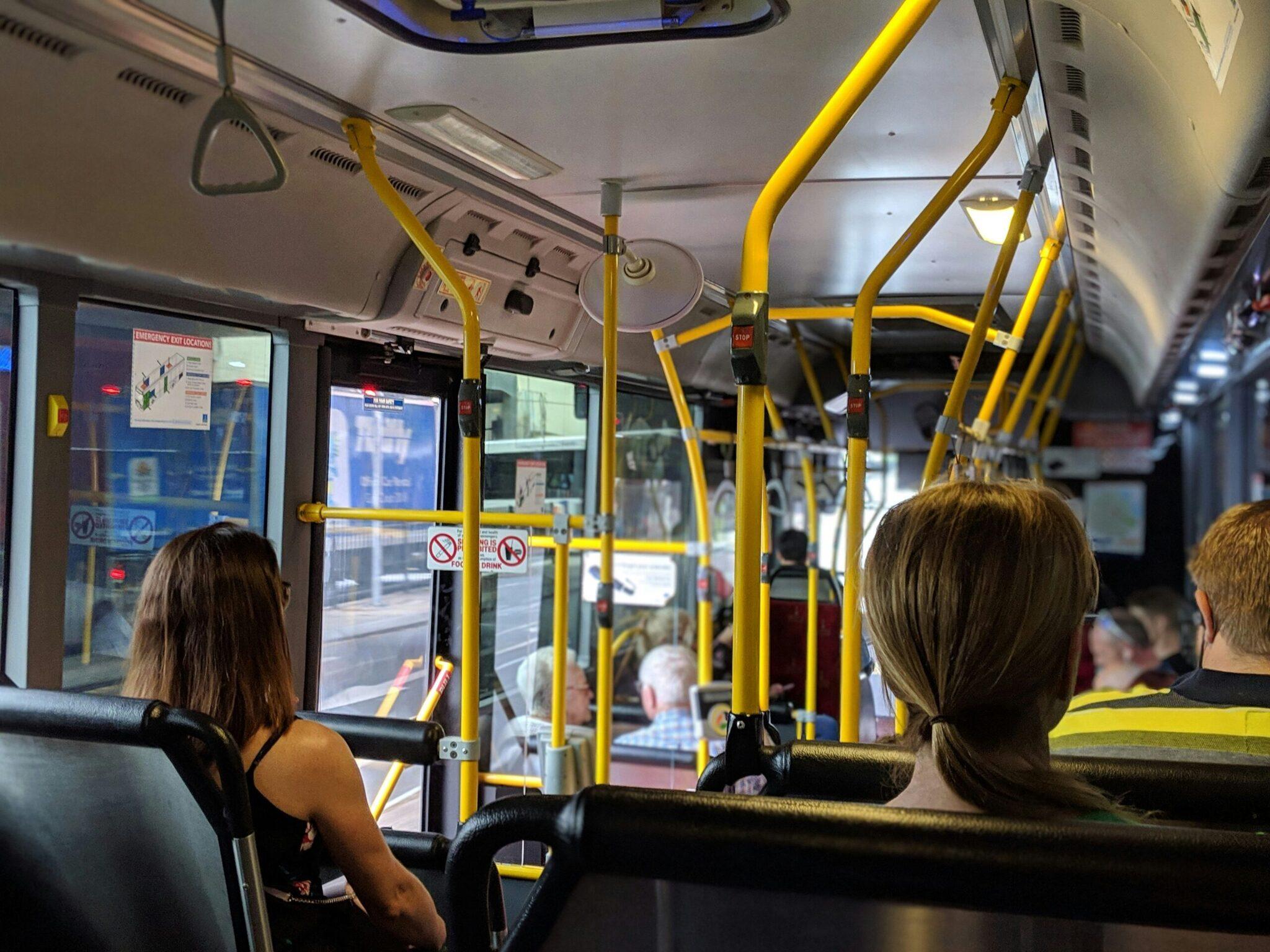 Interior de un autobús con pasajeros sentados, barras de sujeción amarillas y ventanas tintadas con vistas a la calle