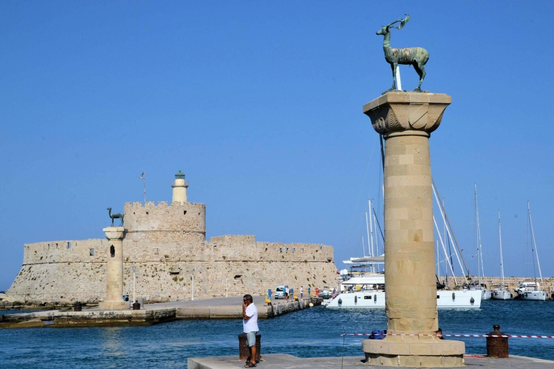 Un porto mediterraneo con colonna sormontata da una statua di cervo, fortezza in pietra e velieri ormeggiati