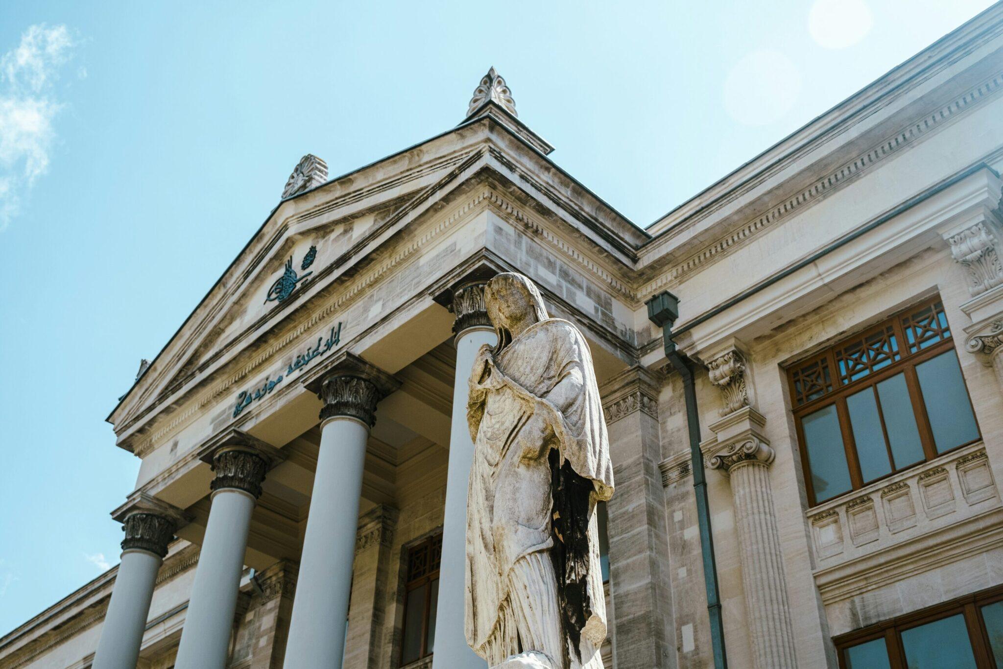 Facciata neoclassica di un museo con colonne corinzie e statua di angelo bianco sotto cielo blu