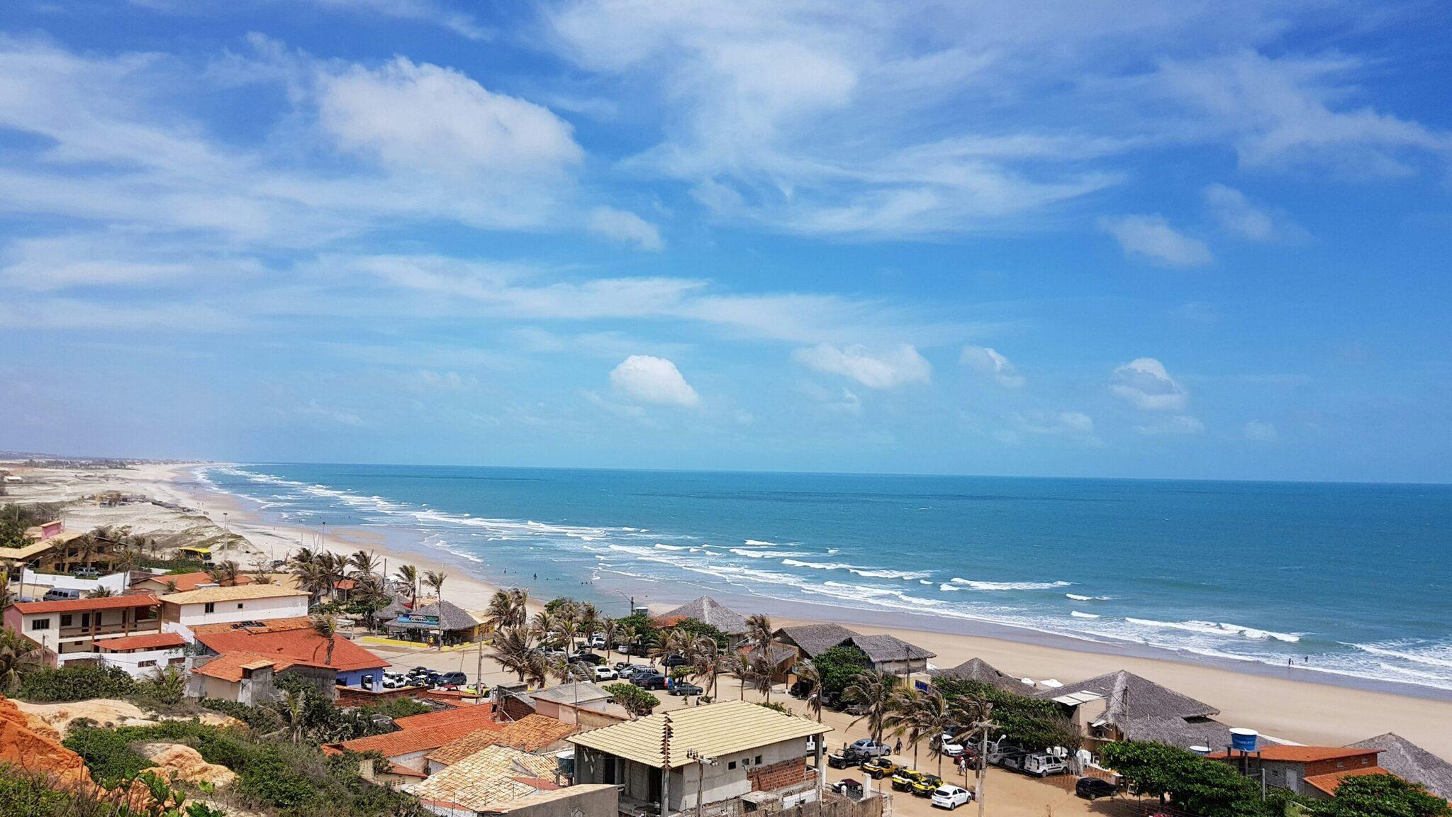 Aerial view of a coastal beach with waves, orange-roofed houses, palm trees and a clear blue sky