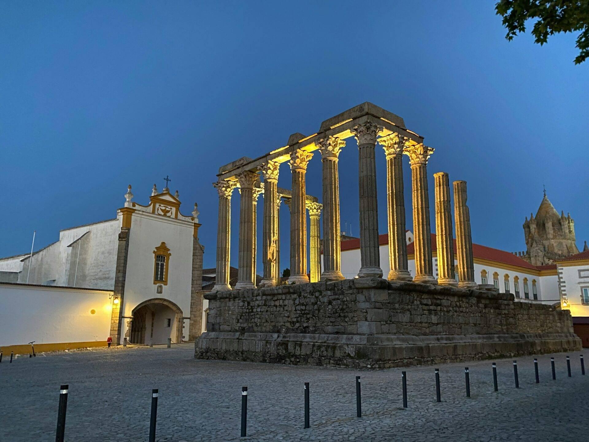 Beleuchtete römische Ruinen mit korinthischen Säulen und weißen Kirchen im Hintergrund in der Abenddämmerung