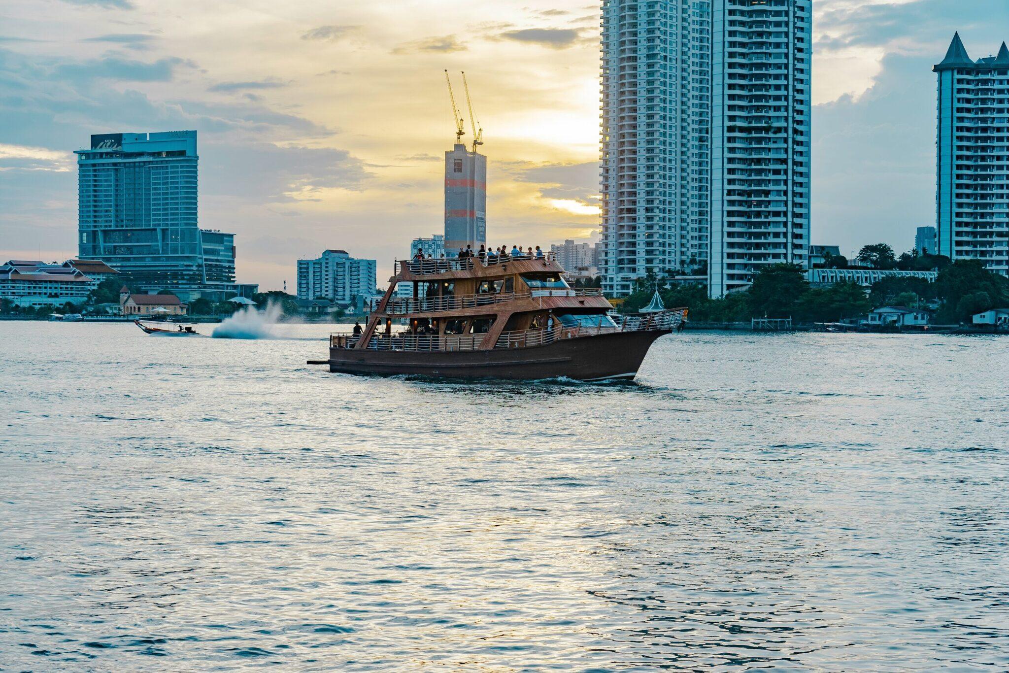 A traditional tour boat cruising a river at sunset with skyscrapers and a temple in the background
