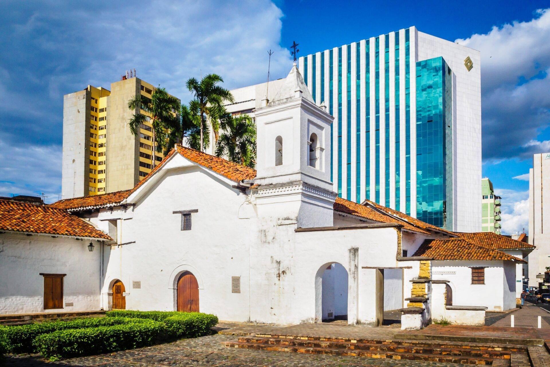 Iglesia colonial blanca con tejados de tejas naranjas que contrasta con edificios modernos al fondo bajo un cielo azul