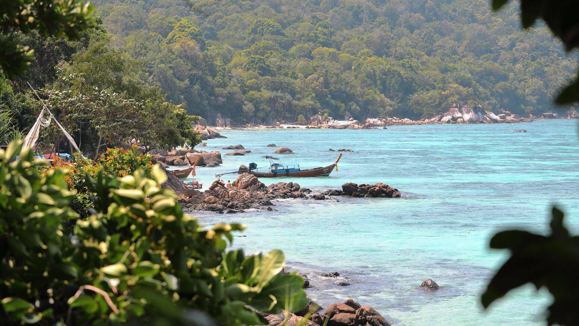 Tropische Küstenansicht mit Longtailboot, türkisblauem Wasser, Felsen und dichtem Dschungel im Hintergrund