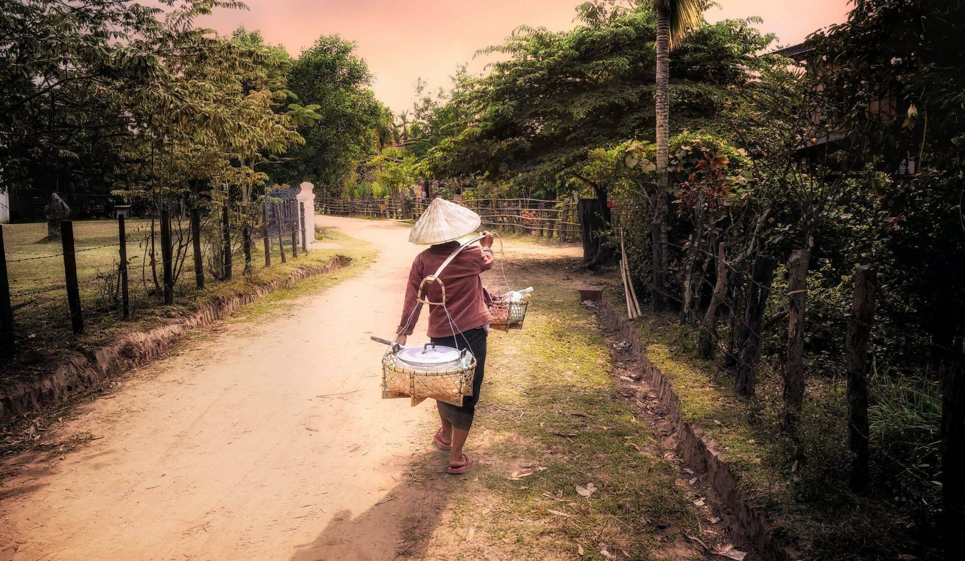 Una donna con cappello conico cammina su un sentiero sterrato bordato di verde nel Laos rurale