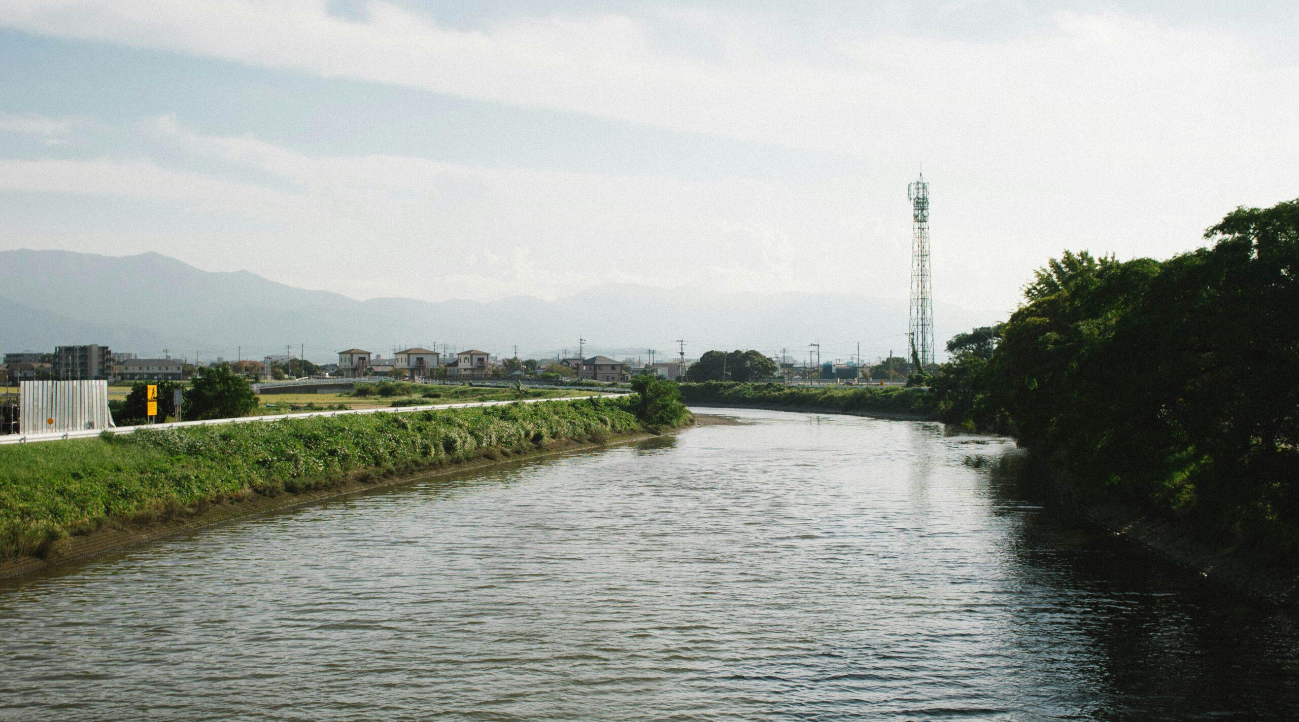 Vista di un fiume che attraversa una zona rurale giapponese con case tradizionali e una torre di comunicazione sullo sfondo