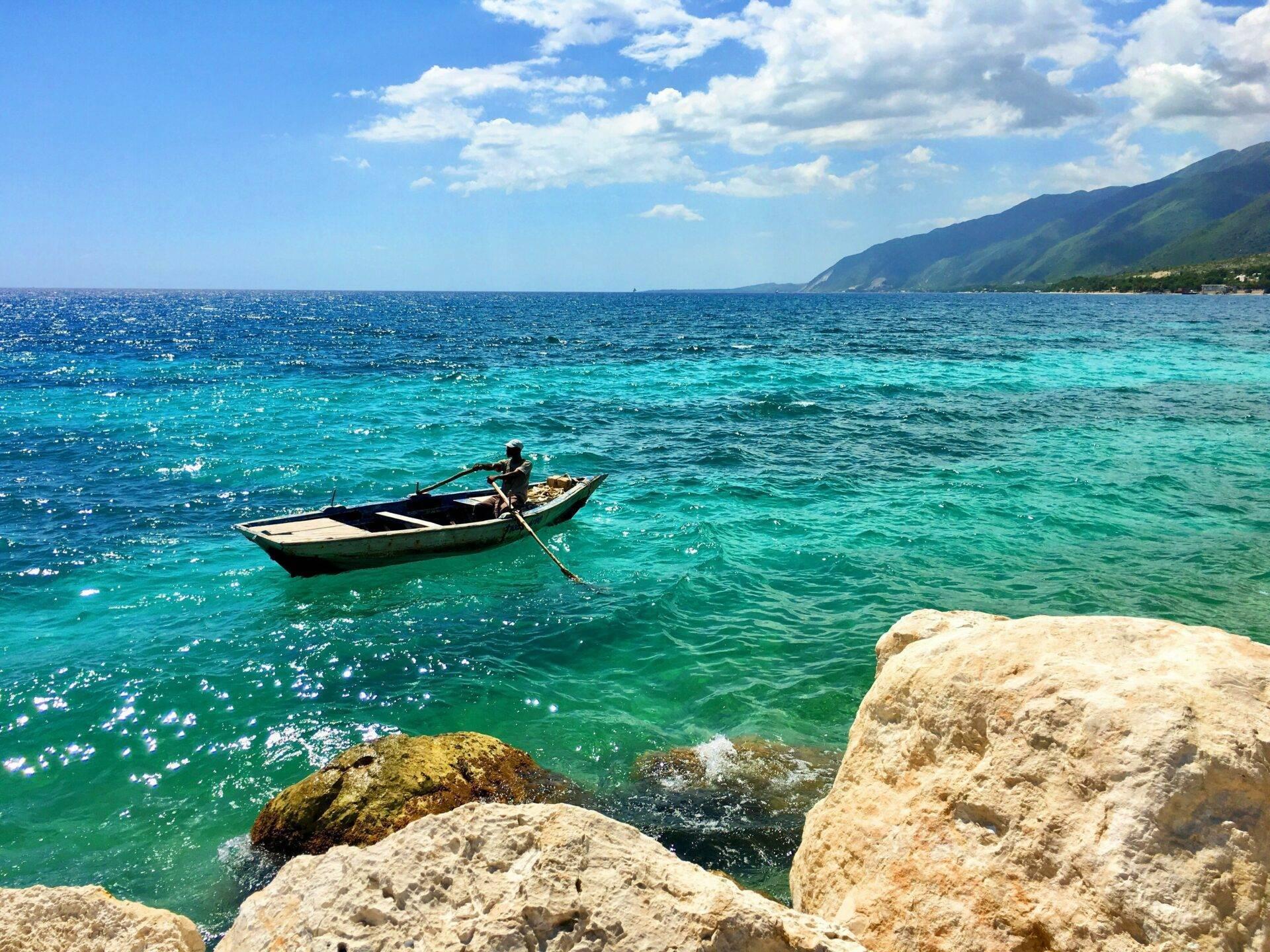 Un pêcheur dans une barque traditionnelle aux eaux turquoise cristallines face à des montagnes verdoyantes côtières