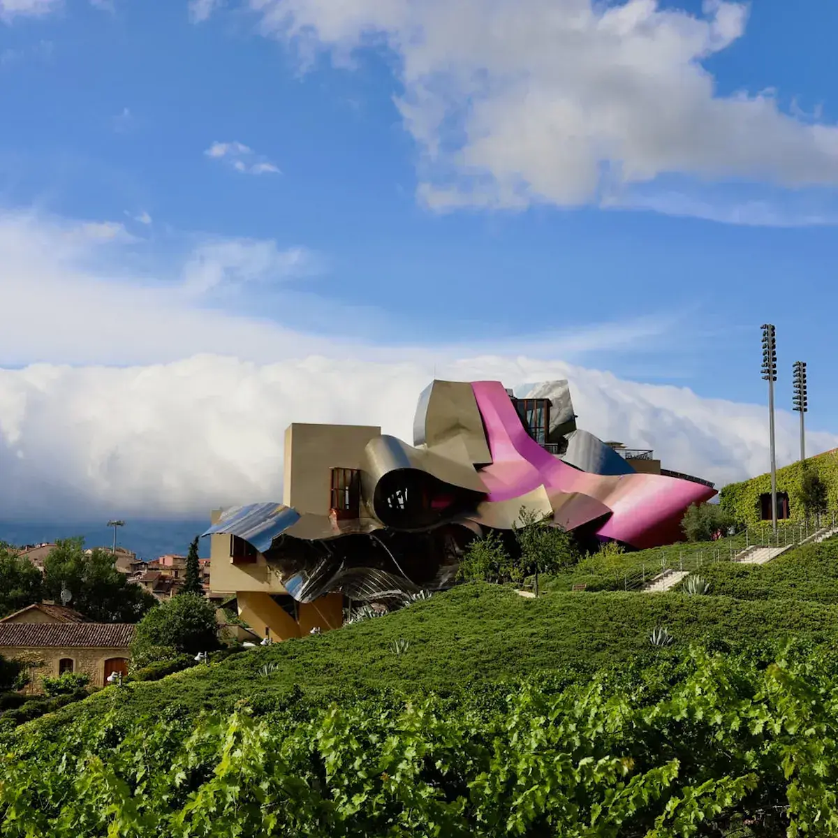 Ein futuristisches Gebäude mit geschwungenen, leuchtend rosa Formen überragt grüne Weinberge in Hanglage