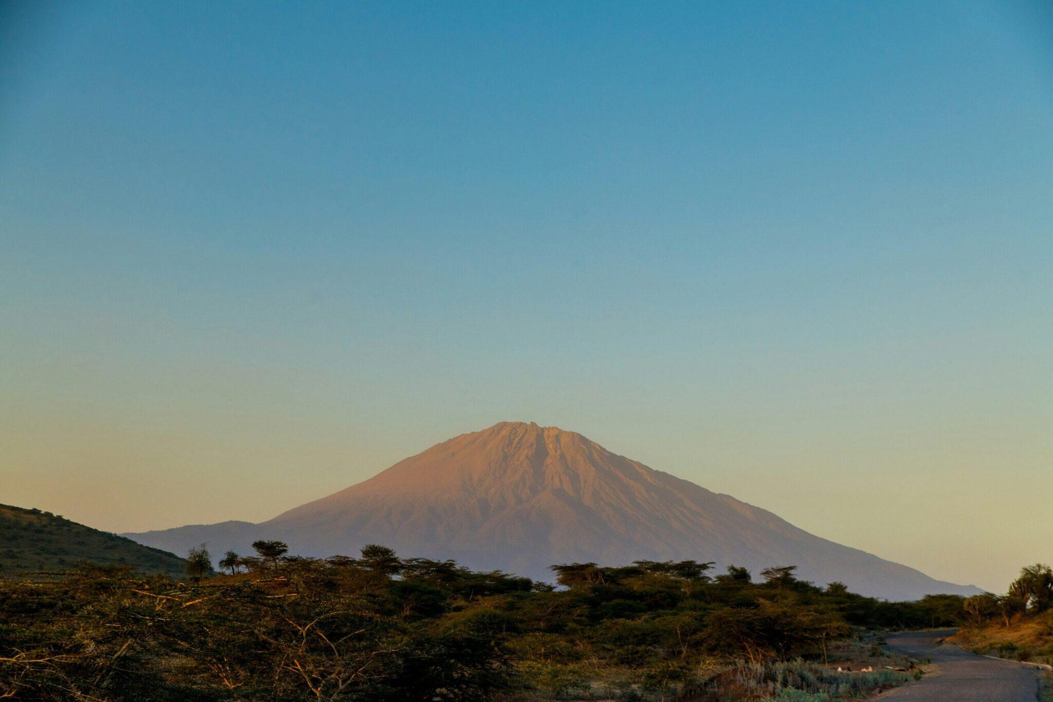 Un volcán majestuoso de laderas estriadas domina un paisaje de sabana árida con acacias dispersas y un río en primer plano