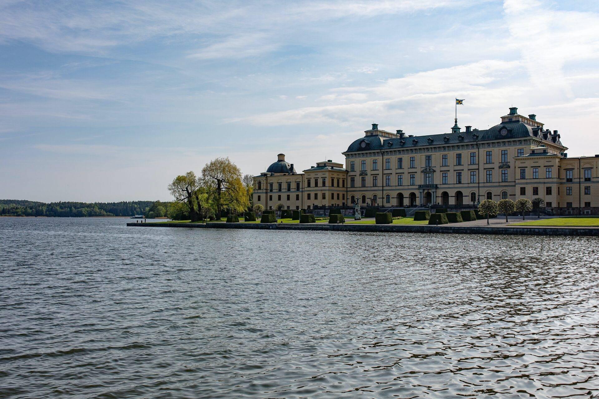 Ein majestätisches gelbes Barockschloss am ruhigen See, umgeben von Grün unter bewölktem Himmel