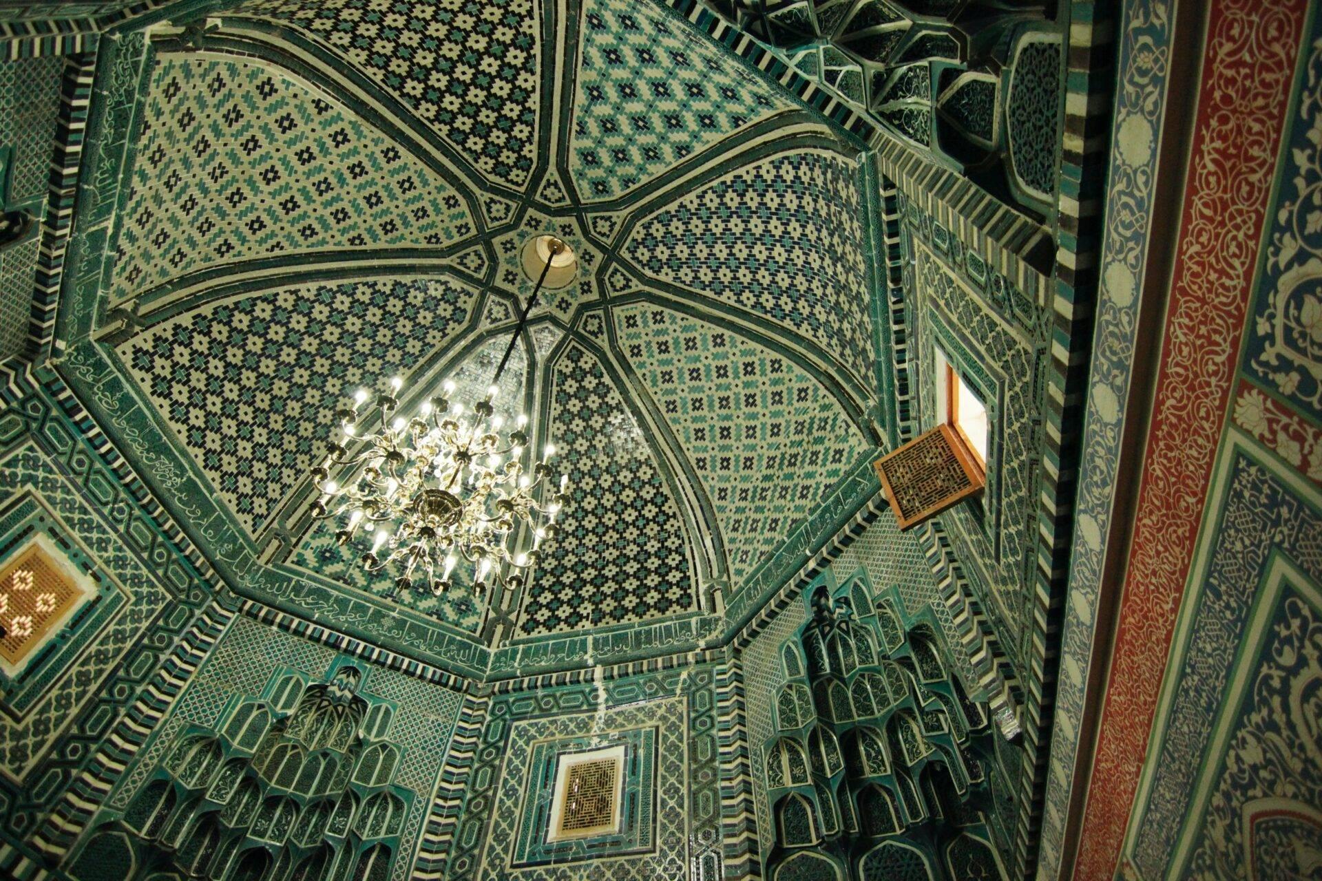 Vogelperspektive auf das Innere einer Moschee mit achteckiger Schmuckdecke, türkisfarbenem Kachelwerk und zentralem Kronleuchter