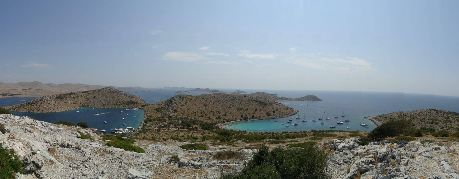 Vista panoramica di una baia mediterranea con isole boscose, acque turchesi e barche a vela all’ancora