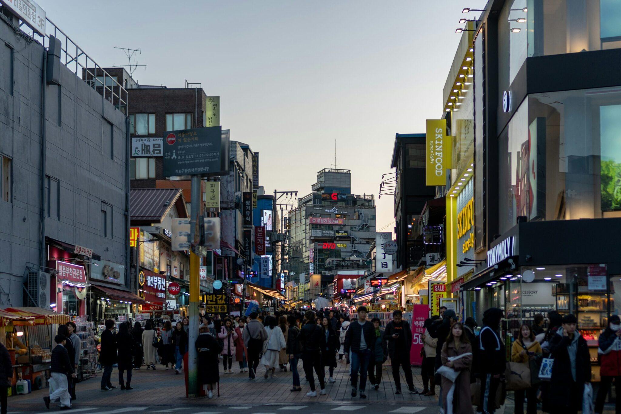Eine belebte Einkaufsstraße in Seoul mit Menschenmassen, koreanischen Leuchtreklamen und Geschäften am frühen Abend