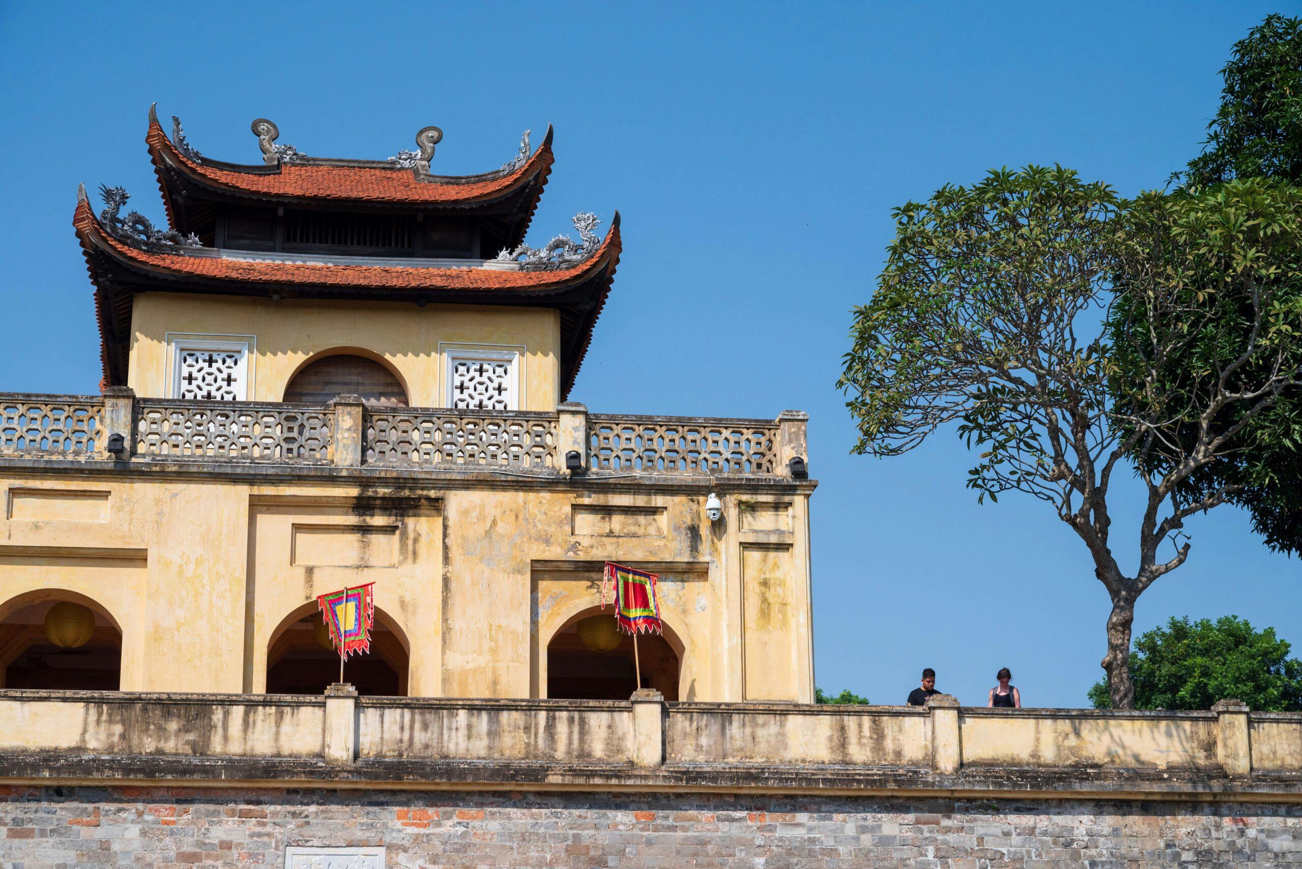 Templo asiático con tejados curvados rojos y paredes amarillas con balcón florido bajo cielo azul
