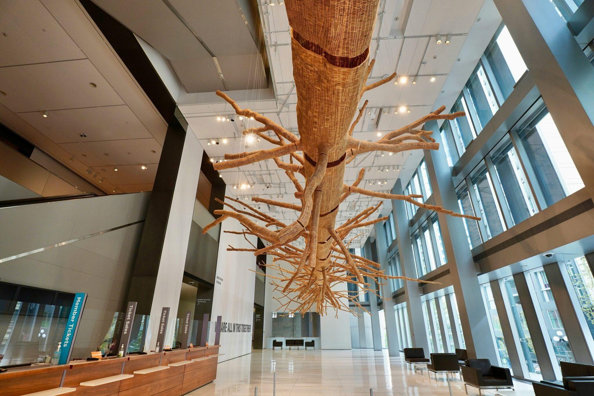 Una instalación artística en madera que representa un árbol gigante suspendido del techo de un luminoso hall moderno