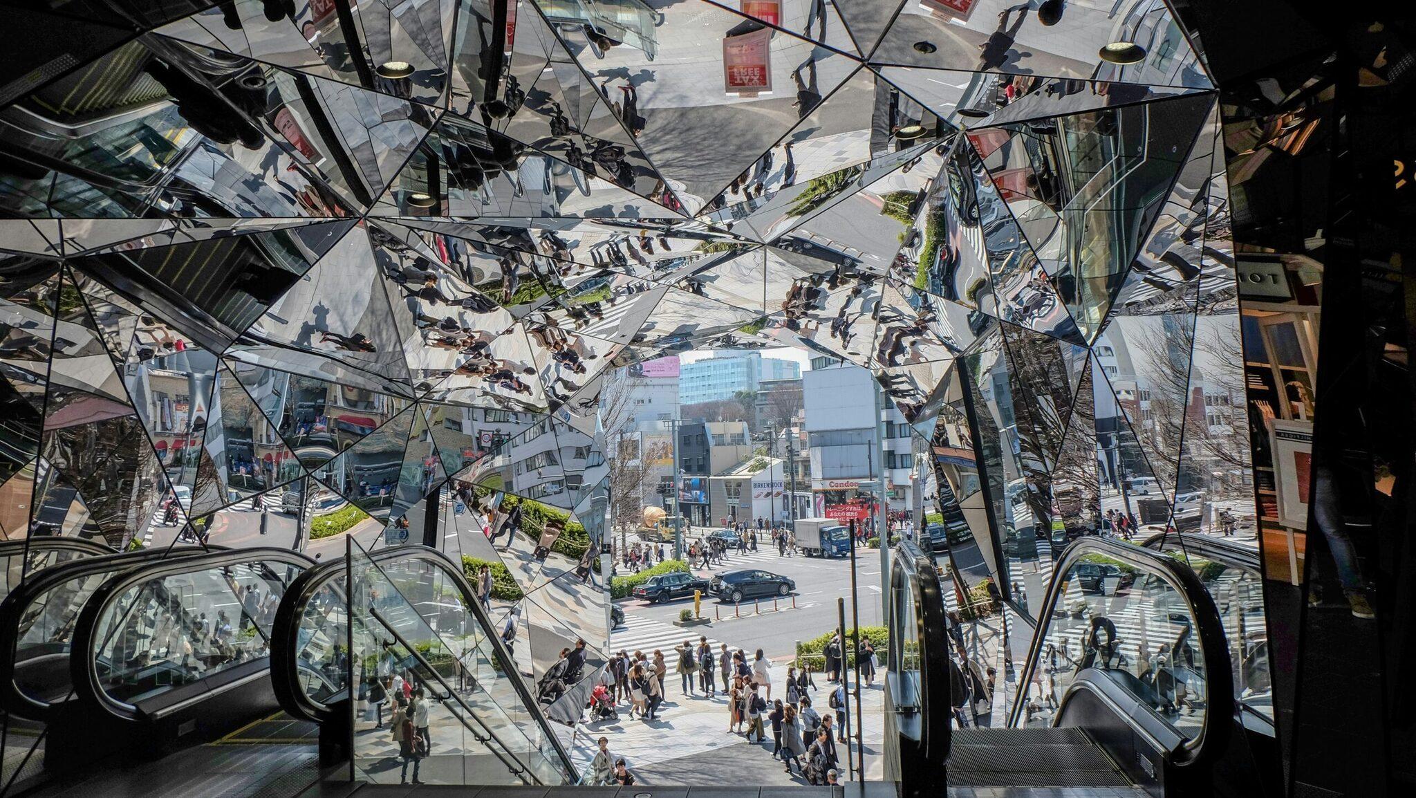 Vista desde el interior de un centro comercial hacia una plaza pública animada con escaleras mecánicas y edificios urbanos