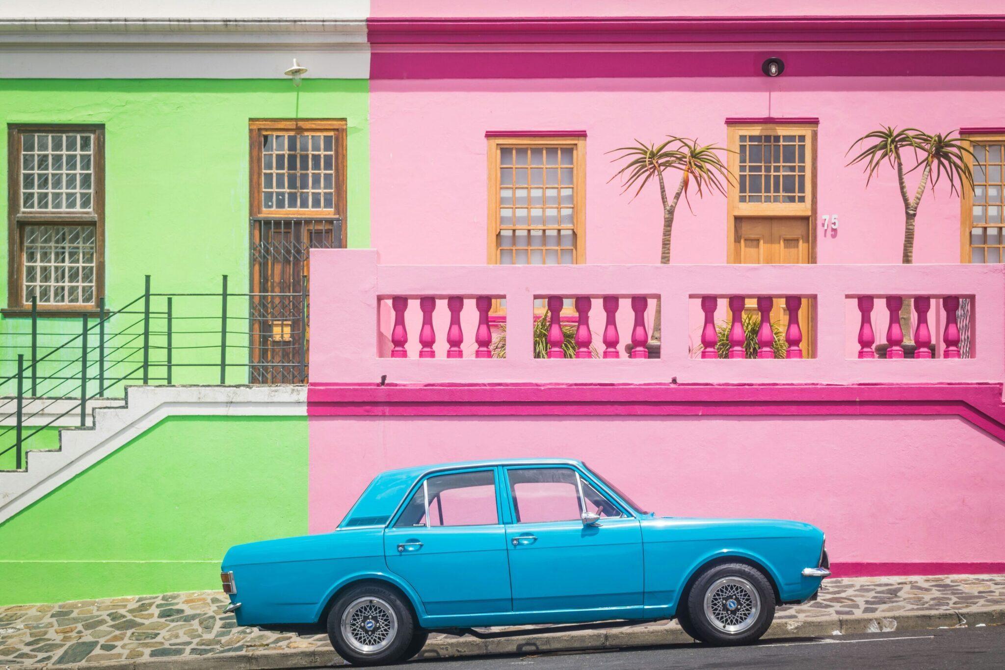 Un’auto blu d’epoca parcheggiata davanti a facciate colorate rosa e verde con balconi ornamentali e palme