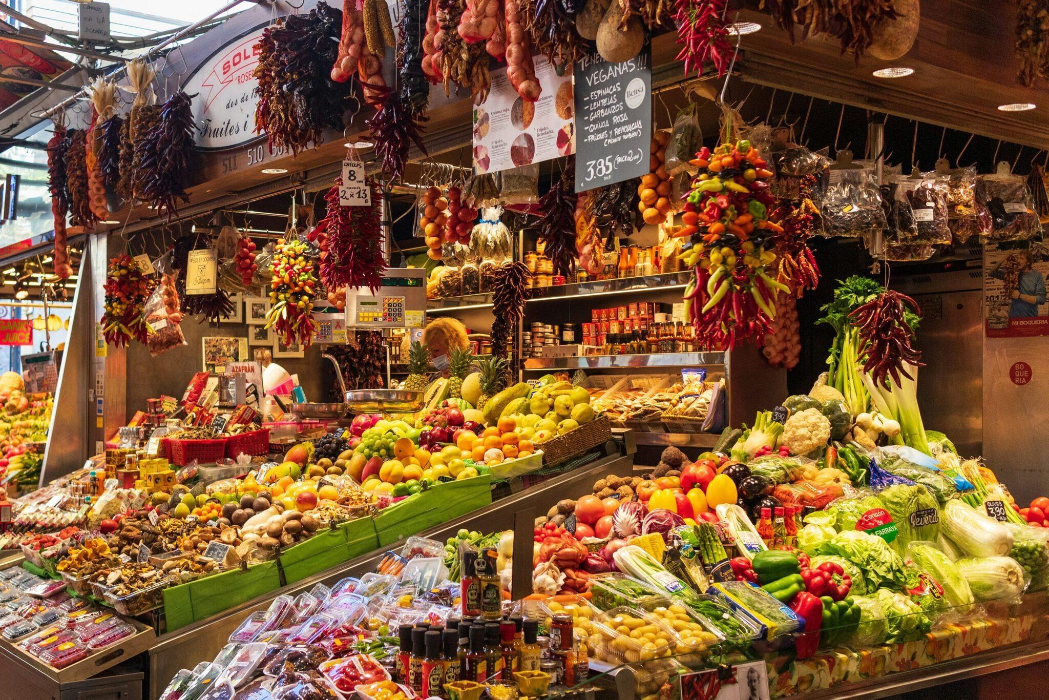 Puesto de mercado cubierto repleto de frutas, verduras frescas y productos locales colgantes, que ilustra la riqueza gastronómica mediterránea
