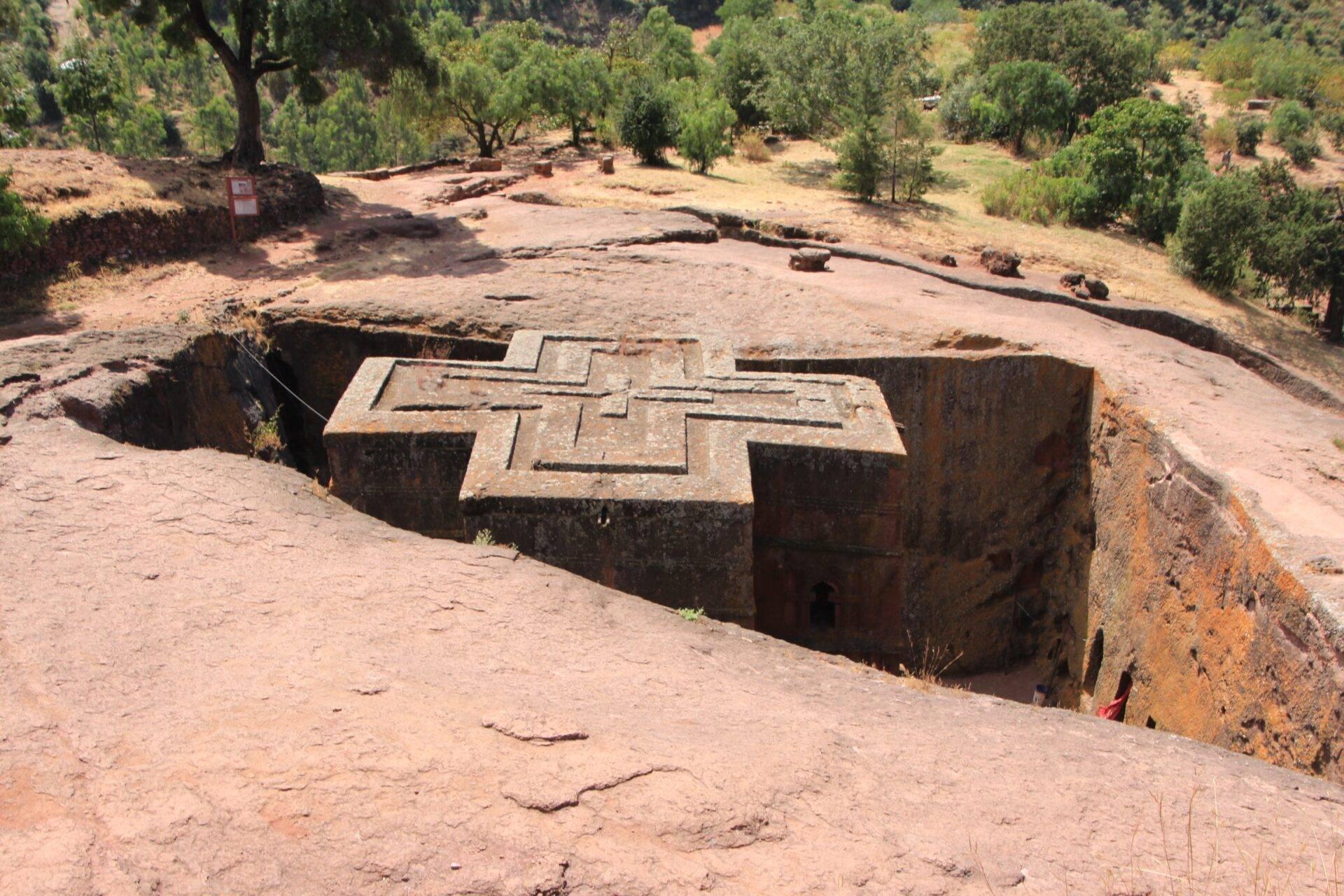 Rock-hewn church carved into a cliff face with a cross cut into the roof, surrounded by trees and arid terrain