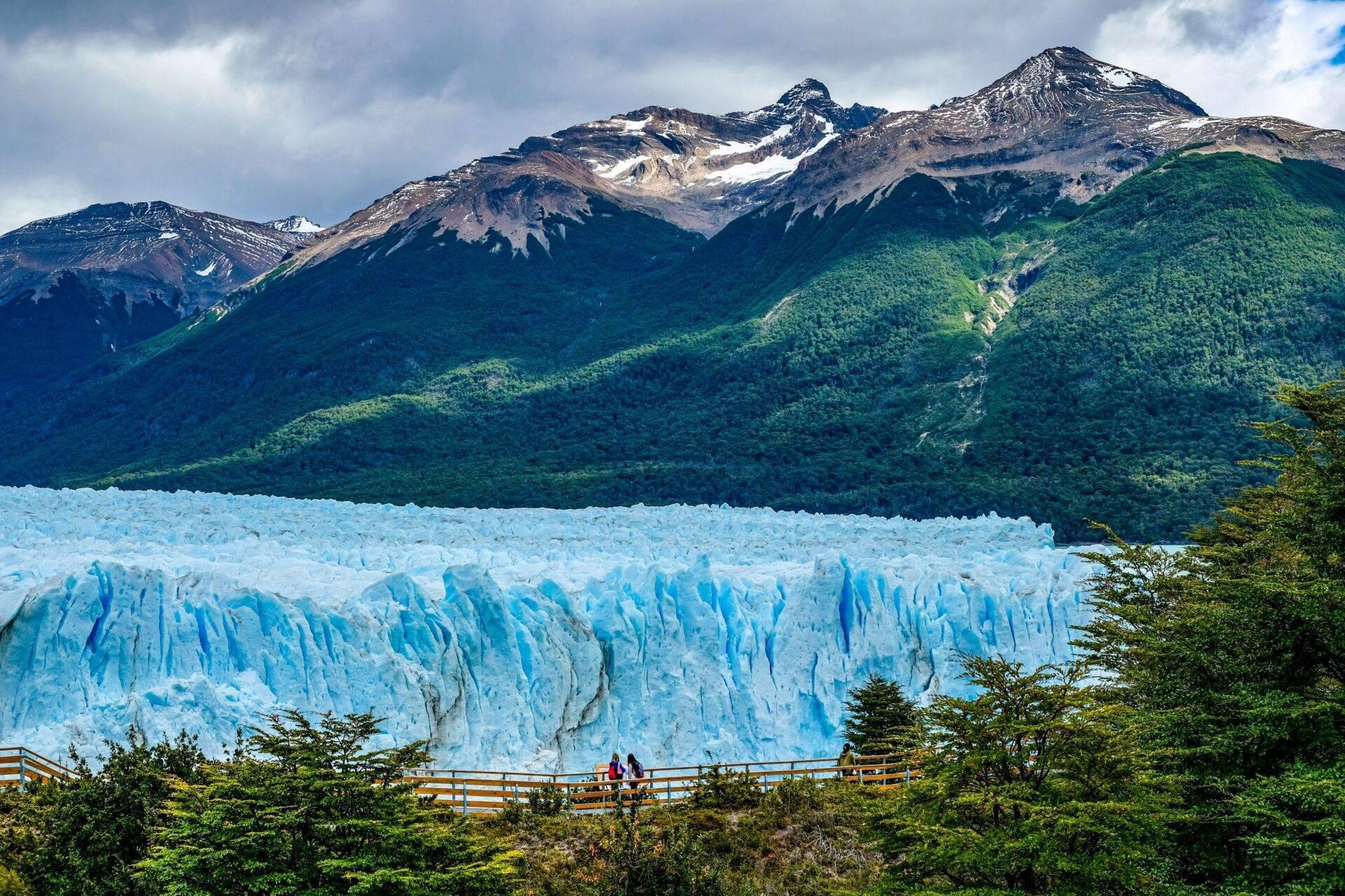 Un imponente ghiacciaio blu di fronte a montagne innevate e una fitta foresta sotto un cielo nuvoloso