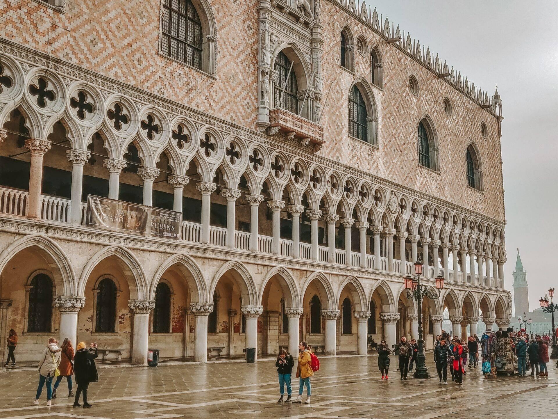 Ornate facade of a Venetian palace with a colonnaded gallery and arched arcades, visitors on the wet square below